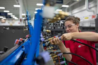 A female student wearing clear safety glasses and a red polo shirt labeled "AMT" (Advanced Manufacturing Technician) works inside a bright, industrial-style training lab. She is connecting a black hose with a metallic fitting to a blue equipment panel that features multiple valves and colored connectors. The background shows rows of machinery bays, overhead lighting, and a clean, organized workshop environment focused on technical skill development.